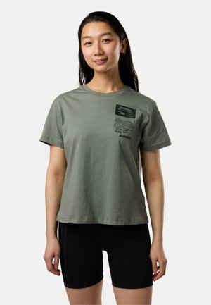 Jeune femme portant un t-shirt vert graphique et un short de cyclisme noir, debout devant un fond uni clair.