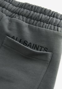 Sive hlače z elastičnim pasom, z žeparjem, ki prikazuje ime blagovne znamke "ALLSAINTS" v črnem tisku.