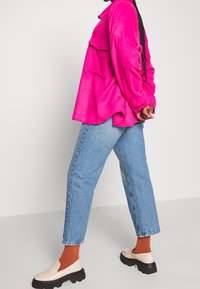 Camisa de manga larga fucsia con bolsillo frontal, combinada con jeans rectos de color azul claro y zapatos sin cordones claros con suela negra gruesa.