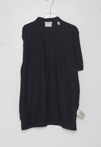 Tricou polo negru cu mâneci scurte atârnat pe un umeraș transparent, etichetă GANT vizibilă, etichetă de mărime medie atașată lângă guler și cusătura laterală.