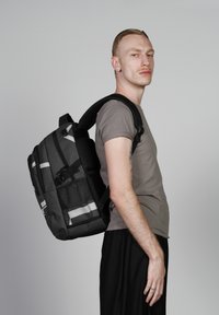 Sac à dos noir avec une surface texturée, doté de bandes réfléchissantes, de plusieurs compartiments et de bretelles rembourrées, porté par un modèle.