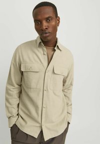 Camicia beige a maniche lunghe con due tasche frontali, colletto alla francese e una finitura testurizzata. Indossata sopra un top marrone chiaro.