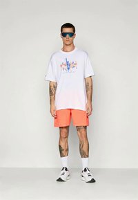 Balta grafiska T-kreklina ar krāsainu druku un korallkrāsas šortiem. Modelis valkā zila saulesbrilles un baltas tenisa kedas. Gluda tekstūra, ikdienišķs piegriezums.