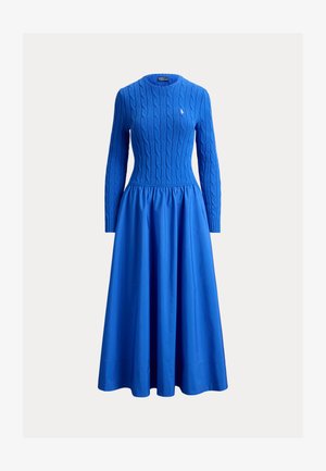 Blauwe jurk met lange mouwen, voorzien van een gebreid lijfje met kabelpatroon en een geplooide stoffen rok. De taille is aangesloten en benadrukt de vorm.