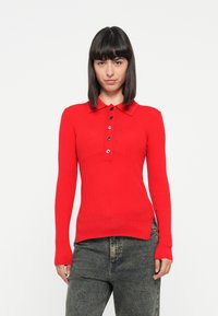 Polo shirt rouge à manches longues et à côtes, avec un col et des boutons noirs, présentant une coupe ajustée et des fentes sur les côtés au niveau de l'ourlet, assorti à un jean sombre.