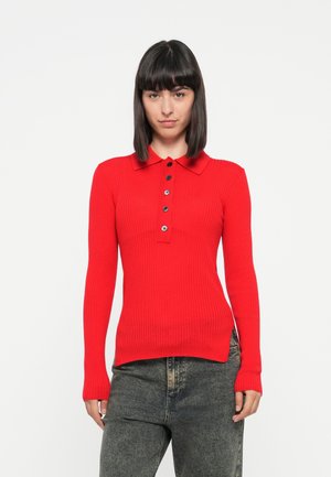 Polo shirt rouge à manches longues et à côtes, avec un col et des boutons noirs, présentant une coupe ajustée et des fentes sur les côtés au niveau de l'ourlet, assorti à un jean sombre.