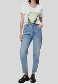 Camiseta blanca con un gráfico de unos auriculares verdes y plateados, combinada con unos vaqueros de mezclilla azul claro de cintura alta y sandalias negras de tiras.