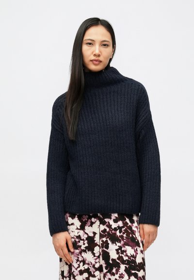 Pull en maille côtelée bleu marine avec un col montant, manches longues et coupe décontractée, porté avec une jupe à motif floral.