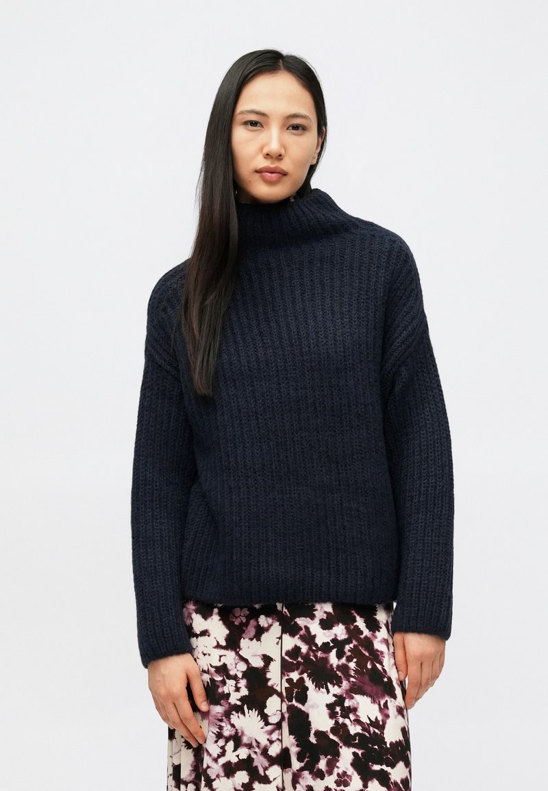 Πλεκτό πουλόβερ σε σκούρο μπλε χρώμα με ριμπ ύφανση και ψηλό λαιμό, με μακριά μανίκια και άνετη εφαρμογή, φορεμένο πάνω από φούστα με φλοράλ μοτίβο.