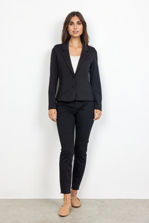 Blazer noir ajusté avec un bouton unique, revers crantés, associé à un pantalon noir slim et des chaussures à enfiler beiges sur un fond neutre.
