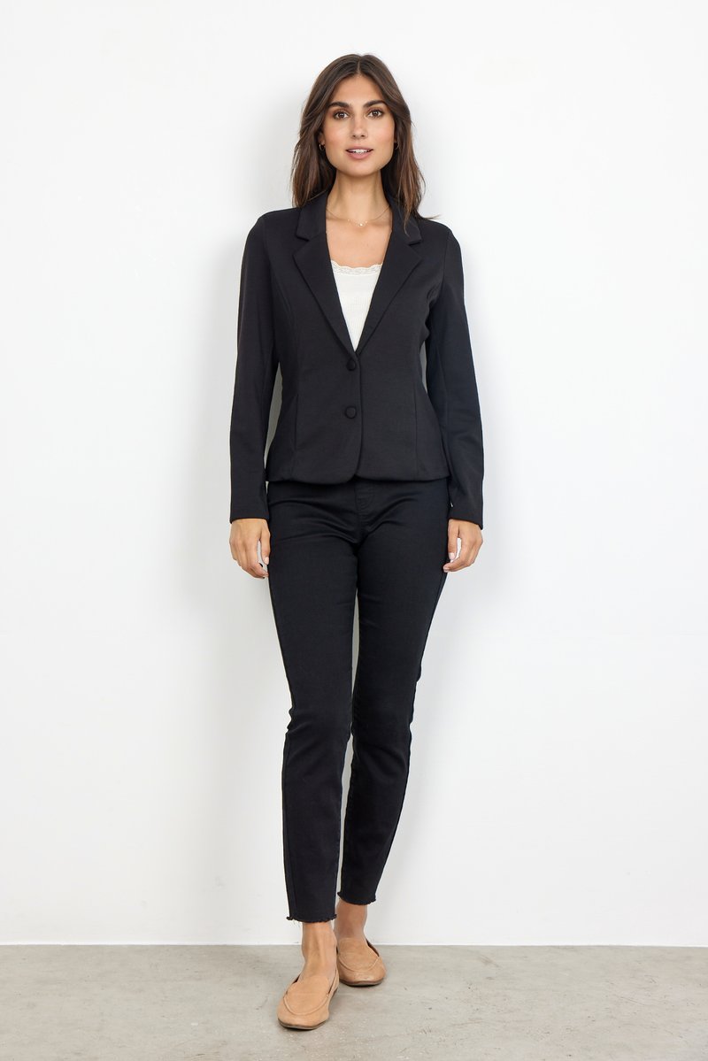 Schwarze, taillierte Blazerjacke mit einem einzigen Knopf, mit Reverskragen, kombiniert mit schwarzen Slim-Fit-Hosen und beigen Slippern vor einem neutralen Hintergrund.
