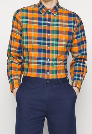 Homme portant une chemise à manches longues boutonnée avec un motif à carreaux orange, bleu, vert et marron rentrée dans un pantalon bleu marine.