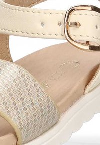 Sandalia beige con una correa texturada que presenta un sutil brillo metálico, una correa para el tobillo con una hebilla dorada y una suela blanca.