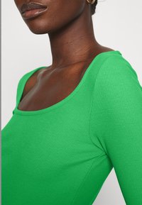 Kvinna i en ljusgrön ribbad långärmad tröja med rundad halsringning och guldringar i öronen mot en enfärgad bakgrund.