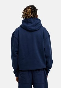 Marineblauer Kapuzenpullover mit lockerer Passform, ausgestattet mit einer großen Kapuze und tiefen Schultern. Das Material sieht weich aus und hat minimale Details.