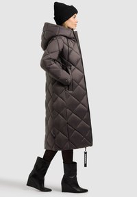 Vrouw in een lange, gewatteerde donkere jas met capuchon, zwarte beanie, zwarte legging en zwarte sleehaklaarzen die tegen een effen achtergrond loopt.