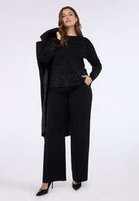 Maglione nero con decorazioni argentate decorative, abbinato a pantaloni neri a gamba larga e tacchi alti neri, cappotto drappeggiato su una spalla.