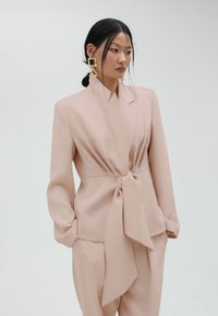 Svijetlo ružičasti sako s čvorastim detaljem sprijeda, dugim rukavima i oštrim kragnom; u kombinaciji s odgovarajućim krojenim hlačama.