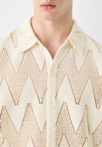 Maglietta a maglia color crema con motivo a zig zag, colletto corto e bottoni frontali, con un design a tessitura aperta e texture.
