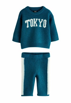 Young boy with blond hair wearing a teal sweater saying "TOKYO" and matching pants with white stripes, standing and gesturing with hands.