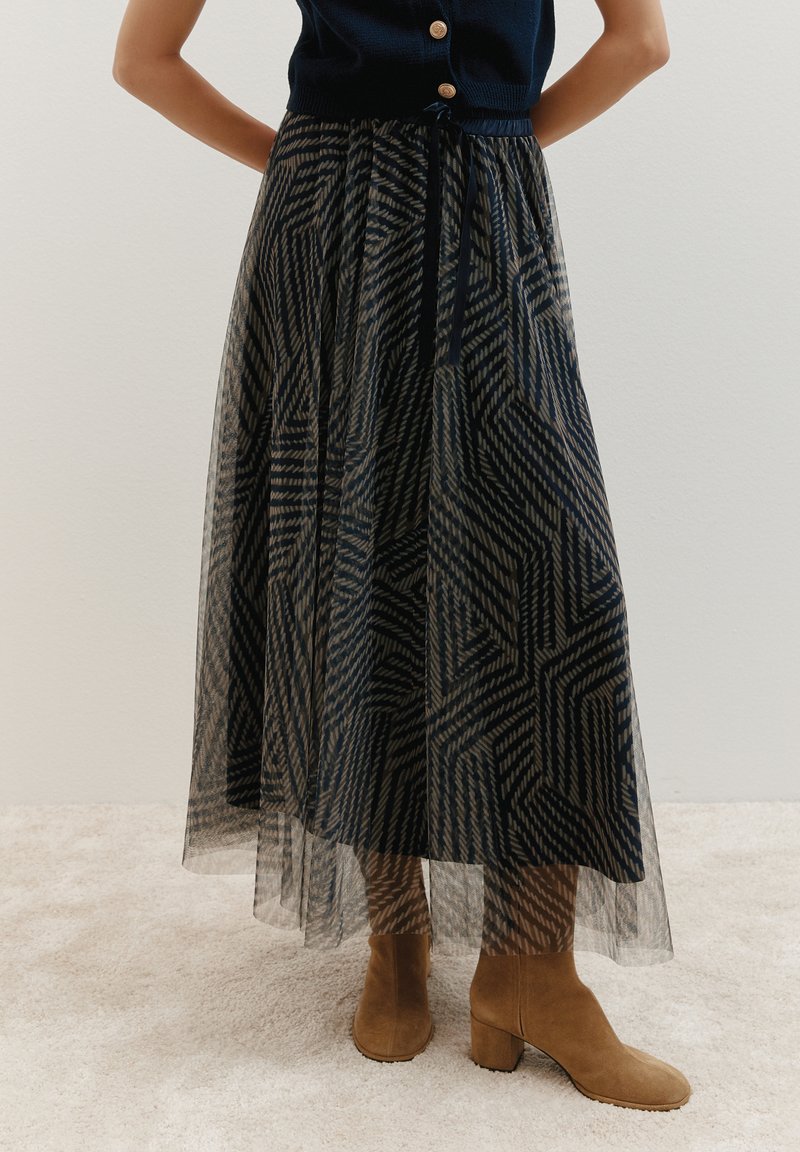 Jupe maxi en motifs géométriques bleu marine et crème, fabriquée en tissu léger et transparent, dotée d'une taille à cordon de serrage et associée à des bottes à talons marron.