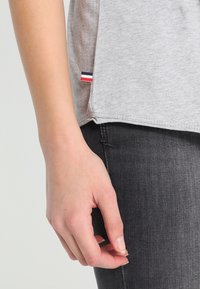 Grå kortärmad skjorta med rundad nederkant och en liten tagg med röd, vit och blå rand på sidan, ihop med mörka jeans.