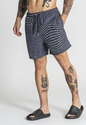 Mand iført marineblå mønstrede shorts med tekstdesign, sorte slidesandaler og flere tatoveringer på ben og arme, stående mod en ensfarvet baggrund.