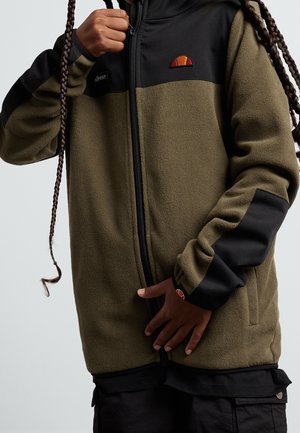 Personne portant une veste polaire zippée verte et noire avec un logo sur la poitrine et la manche, tenant la zone de la fermeture éclair avec les deux mains.