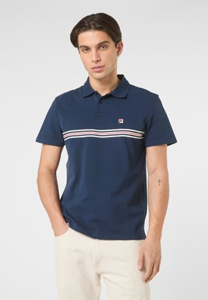 Mand iført marineblå polo skjorte med hvide, røde og blå vandrette striber, stående med den ene hånd i lommen mod en ensfarvet baggrund.