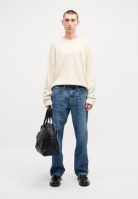 Κρέμα oversized πουλόβερ με κεντημένο λογότυπο, σε συνδυασμό με ανοιχτό μπλε τζιν ίσιας γραμμής. Το μοντέλο κρατά μια μαύρη δερμάτινη τσάντα και φορά μαύρα παπούτσια.