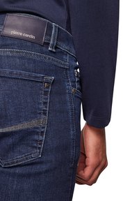 Jean en denim bleu foncé avec une étiquette en cuir portant la mention "Pierre Cardin". Les caractéristiques incluent une poche arrière avec un détail de couture et une coupe slim.
