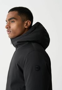 Juodas sukapotas džemperis, pagamintas iš lygaus, vandeniui atsparaus audinio. Funkcija - užtrauktuko uždarymas ir apvalus logotipo pleistras kairiajame rankove.