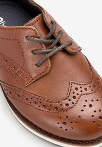 Brązowe skórzane buty garniturowe z perforowanymi detalami, szarymi sznurowadłami i okrągłym noskiem. Posiadają kontrastującą białą podeszwę i gładką teksturę.