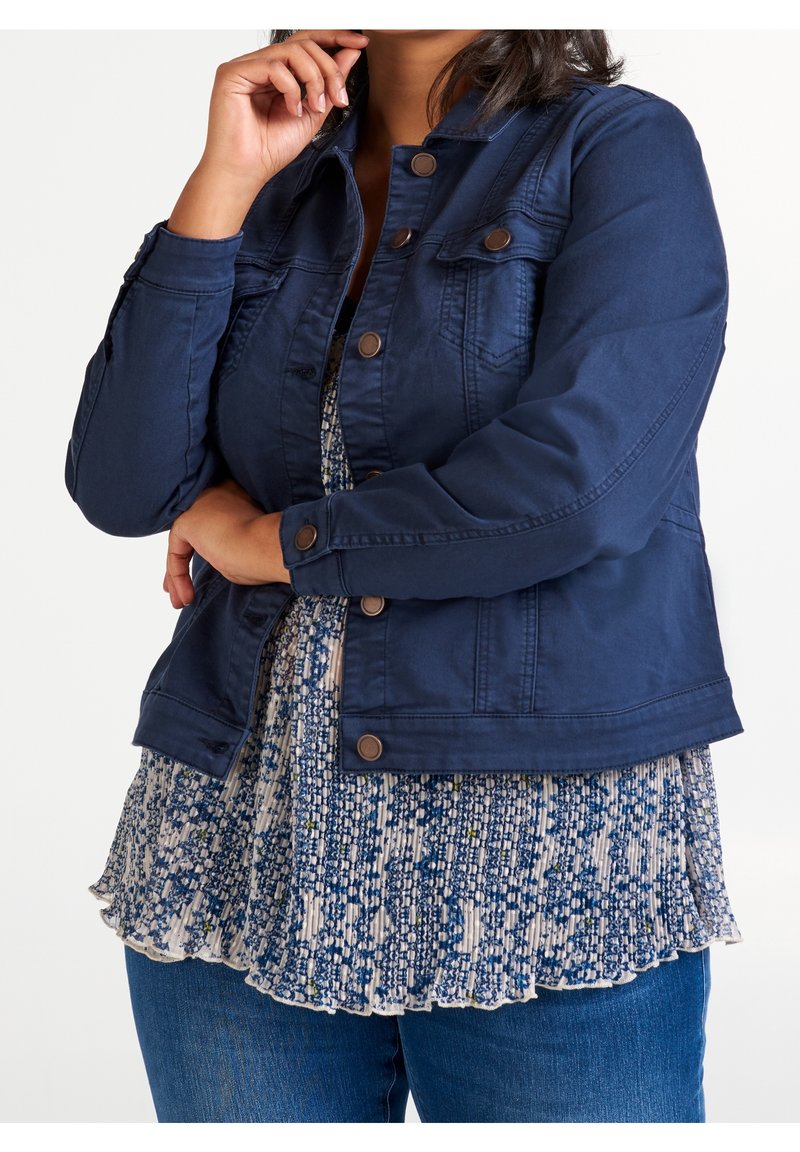 Mujer vestida con una chaqueta de denim azul oscuro abotonada, blusa plisada azul con un patrón y jeans azules, con los brazos cruzados y una mano cerca de la cara.