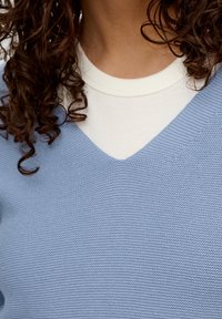 Lichtblauwe gebreide trui met een V-halsontwerp, gedragen over een witte ronde hals T-shirt. De textuur is geribbeld, zichtbaar bij de halslijn.