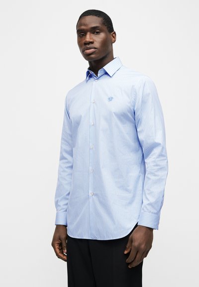 Νεαρός άνδρας που φοράει ανοιχτό μπλε και λευκό ριγέ πουκάμισο με κεντητό λογότυπο στο στήθος, στέκεται μπροστά σε ένα απλό φόντο.