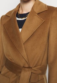 Abrigo de lana marrón con cinturón y grandes solapas, llevado sobre un suéter a rayas azul y blanco y un collar de cadena dorada.