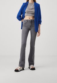 Blazer bleu, haut plissé gris court, jeans gris évasés, et sandales noires avec des lanières blanches et des accessoires argentés.