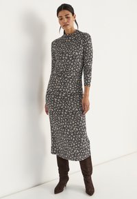 Zwart langemouwen jurk met een bloemenpatroon in crème en roze, voorzien van een hoge halslijn en een getailleerde silhoutte, gecombineerd met bruin kniehoogte laarzen.
