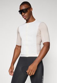 Kortærmet cykeltrøje i beige og hvid med en frontlynlås, strømlinet pasform og åndbart stof. Sorte cykelshorts fuldender looket.