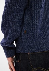 Πλεκτό πουλόβερ σε ναυτικό μπλε χρώμα με ριμπ ύφανση, που διαθέτει μια μικρή πορτοκαλί ετικέτα "BOSS" στο πλάι, συνδυασμένο με σκούρο τζιν παντελόνι.