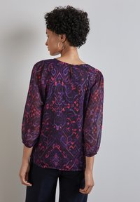 Blusa a righe viola e rossa con maniche a tre quarti, caratterizzata da una chiusura a fessura sul retro e un tessuto leggero e strutturato.