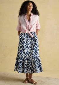 Rozā lina krekls ar ruffu detaļām, sasiets jostasvietā, kopā ar ceļa garuma zilo un balto rakstu svārku, kas rotāts ar flounce apakšmalu.