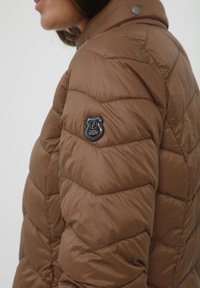 Brun polstret jakke med quilted sektion detaljer. Har en logo patch på venstre arm og en knappet krave. Glat, skinnende tekstur.