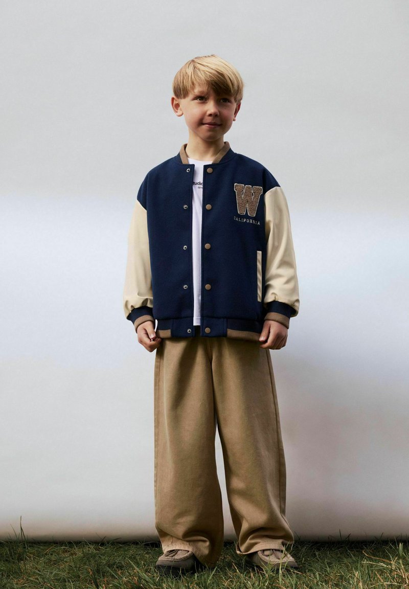 Veste de varsity bleu marine et beige avec un grand patch "W", associée à un pantalon large beige. La tenue comprend un t-shirt blanc en dessous.