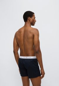 Μαύρο μαγιό σορτς με λευκή μέση που φέρει το λογότυπο "TOMMY HILFIGER". Το μοντέλο εμφανίζεται από πίσω με ουδέτερο φόντο.