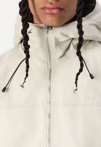 Light beige jacket with a high collar, silver zipper, adjustable drawstring hood, and "OBEY" logo on the chest. Smooth texture.