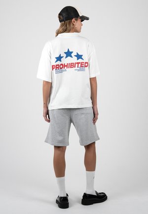 Persona con camiseta blanca de gran tamaño con estrellas azules y texto rojo "PROHIBITED" en la espalda, pantalones cortos grises, zapatos negros, calcetines blancos y gorra negra.