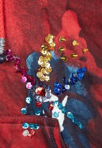 Rød stoftrøje med farverige perler i forskellige nuancer. Designet inkluderer metalliske detaljer og en struktureret overflade.