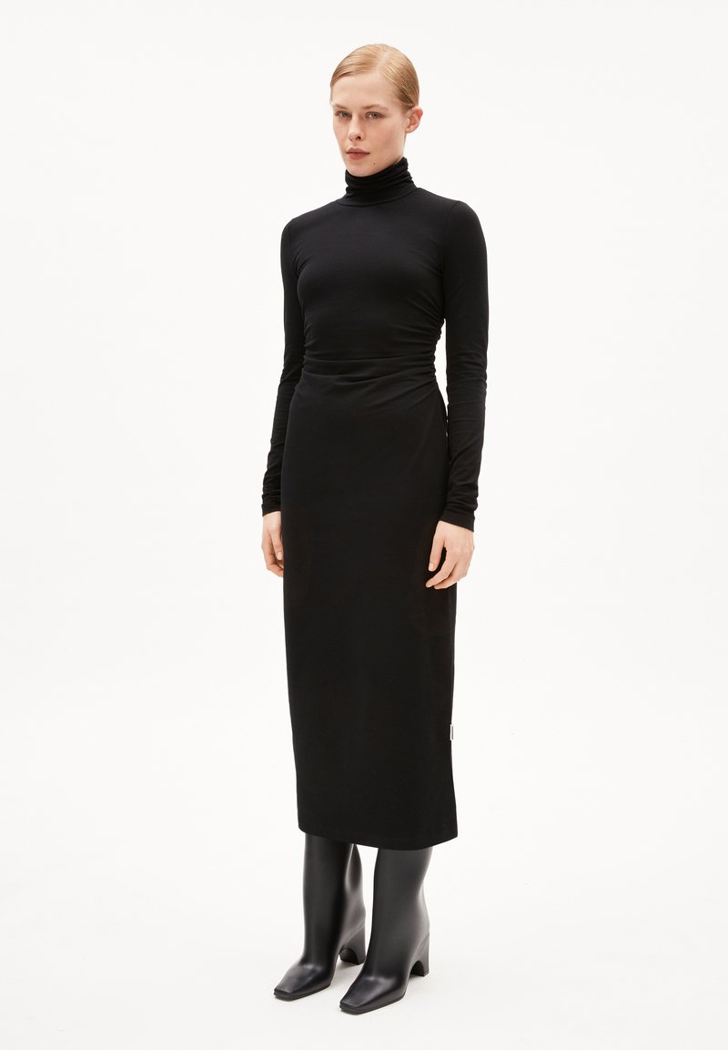 Robe noire ajustée avec un col roulé haut et des manches longues, présentant une texture lisse et élégante ainsi qu'une silhouette droite. Bottes à talons hauts noires.